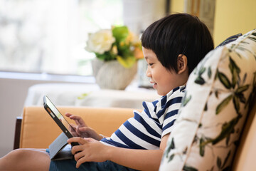 Asian boy is sitting on sofa using a tablet.