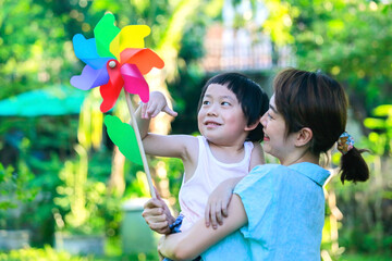 Asian mother is smiling and holding her son trying to spin a colorful wind flower toy over a green garden background.
