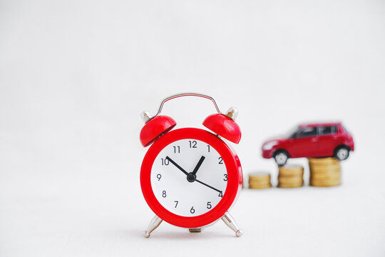 close up of analog red clock with blurred miniature red car on stack of golden coins on grunge light grey background, new car, lower price, sale automobile, reduced price concept