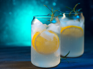 Two glasses of cold summer lemonade with lemon on the rocks and a sprig of rosemary. Colorful blue background, creative stylish glasses