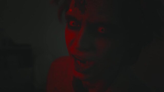 Close up shot of creepy zombie man with white contact lenses and SFX makeup twitching and grunting in darkness with red and blue lights flashing