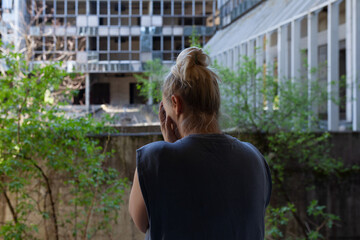 Fototapeta premium Blonde woman standing in front abandoned destroyed building, covering face with hands crying 