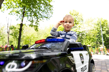 Portrait of cute little caucasain blond toddler boy enjoy having fun riding electric powered police toy car by asphalt path road city park at summer day. Happy child give salute sit vehicle outdoors