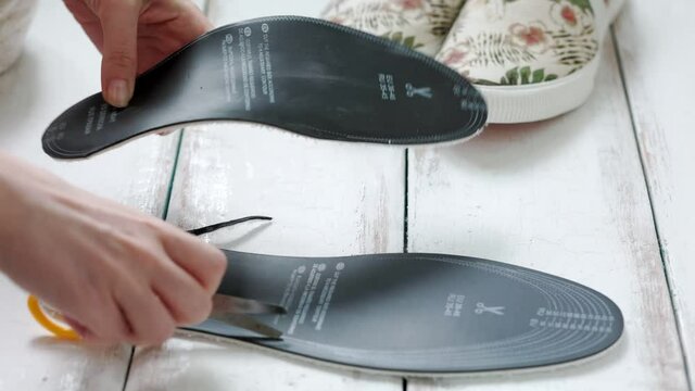 Female's hands cut out a universal orthopedic shoe insole with scissors and insert it into the slip-on. Side view. Close up.