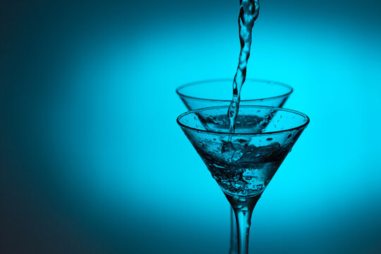 Studio photo of two triangular shape glass and pouring pure water. Transparent martini glass on light blue background. Alcohol or healthy water balance concept.