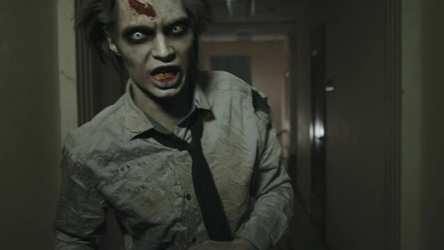 Tracking shot of creepy zombie man with SFX makeup and contact lenses running along dark hallway and following camera