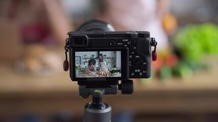 Video camera screen with Asian woman preparing ingredients for healthful vegetarian salad as African American blogger searching recipe online on laptop. Friends recording healthy food blog in kitchen