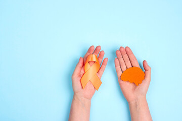 Female hands hold an orange awareness ribbon and brain symbol on a blue background. World Multiple Sclerosis Day.