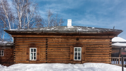 An old one-storey house made of unpainted logs. There are shutters on the windows, a chimney on the roof. There are snowdrifts on the ground. Bare branches and trunks of birches against the blue sky.