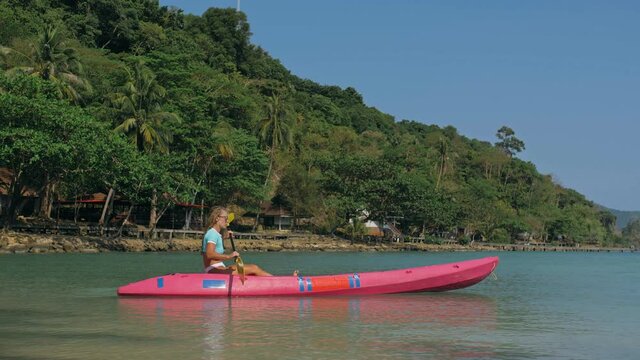 Young blonde woman in blue swimsuit rows pink plastic canoe along azure sea bay past island with palms under blue sky at resort. Traveling to tropical countries. Girl is sailing on kayak in ocean.