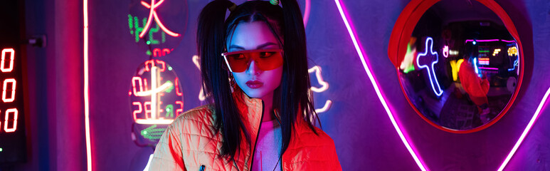 Obraz premium blue lighting on young asian woman in sunglasses posing near mirror and neon lighting, banner