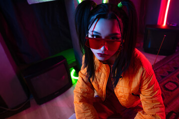 high angle view of stylish young asian woman in sunglasses and orange jacket posing near neon...