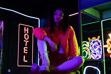brunette young asian woman sitting near neon sign with hotel lettering outside