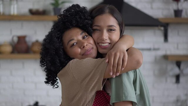 Live camera approaches to happy smiling African American and Asian women hugging in kitchen. Beautiful slim young friends in aprons posing cooking indoors. Friendship and lifestyle