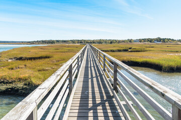 Straight fenced wooden wakway through a grassy coastal marsh on a clear autumn day. Converging lines. Cape Cod, MA, USA.