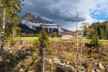 Fotobehang Wooden huts in a meadow at the foot of snowy rocky peaks in the Alps with stom clouds looming on a spring day. A creek is visible in foreground. © alpegor