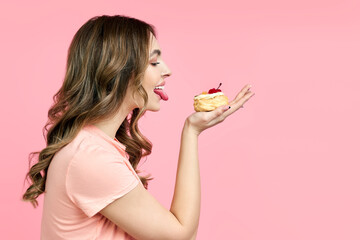 Profile portrait of young woman licking delicious pastry cake on pink background