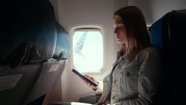 Young Woman Reads E-book while Flying in Airplane and Looking at Window. Touristic Leisure with E-reader or Tablet during Aircraft Flight in Economy Class. 4K Handheld Medium Shot