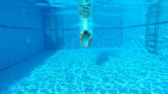 Sporty man diving in pool with crystal clear water. Young guy swimming under water at basin of hotel. Mail tourist relaxing on tropical resort during vacation. Slow motion