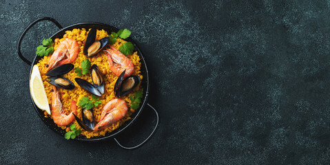 Traditional spanish seafood paella in pan with chickpeas, shrimps, mussels, squid on black concrete background. Top view with copy space. Banner