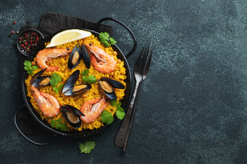 Traditional spanish seafood paella in pan with chickpeas, shrimps, mussels, squid on black concrete background. Top view with copy space - obrazy, fototapety, plakaty