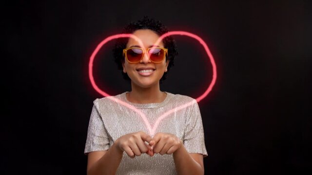 leisure and people concept - african american woman in sunglasses dancing and drawing heart in air with fingers over black background with animated glowing neon lines