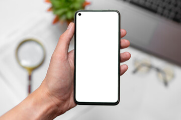 Hand holds smartphone with blank white screen on desktop background, laptop, magnifying glass, houseplant, documents on office table. Layout for design. copy space for text. mockup. top view. 
