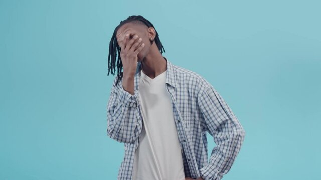 Black frustrated man makes a facepalm gesture and looks discontentedly at the camera on the blue studio background. Black man puts hand on face. Concept of shame and irritation.