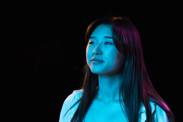 Asian young woman's portrait on dark studio background in neon. Concept of human emotions, facial expression, youth, sales, ad.