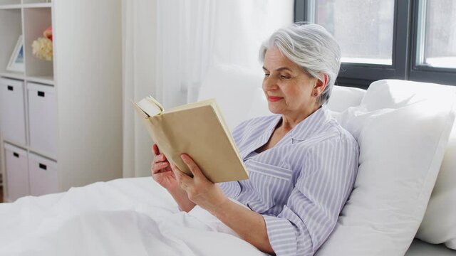 technology, old age and people concept - senior woman reading book in bed at home bedroom