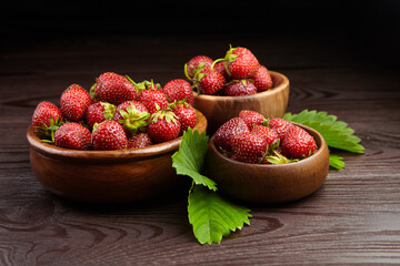 Strawberry red berries in bowls with green leaves on wooden table on black background