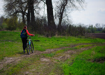 Fototapeta premium a girl with a bicycle walks a field trail. Woman riding a mountain bike in the forest. Hiker woman with backpack. one young woman riding a bike in nature. rolling the bike in his hands. back view.
