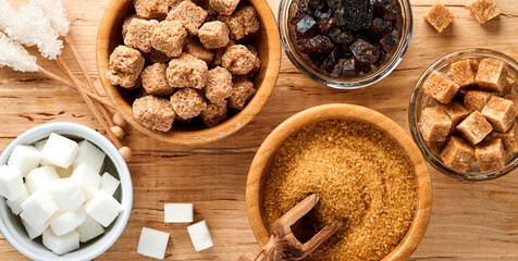 Fototapeta premium White sugar, cane sugar cubes, caramel in bamboo bowl on dark brown table concrete background. Assorted different types of sugar. Top view or flat lay.