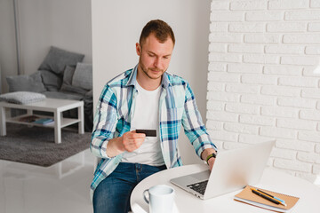 man in shirt sitting in kitchen at home at table working online on laptop