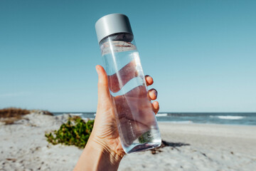 Female hand holding drinking water bottle outdoor on sea shore. Health care concept for water...