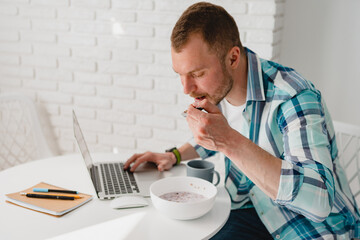 man in shirt sitting in kitchen at home at table working online on laptop