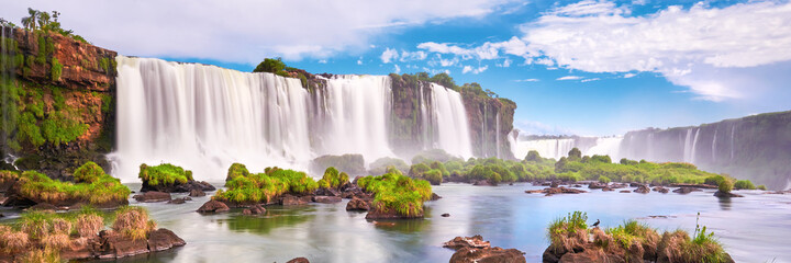 Iguazu waterfalls in Argentina. Panoramic view of many majestic powerful water cascades with mist and clouds. Panoramic image of Iguazu valley with grass and stones in calm water. - obrazy, fototapety, plakaty