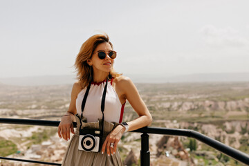 Attractive pretty girl with flying hair wearing sunglasses and holding retro camera looking away and smiling on background of mountains 