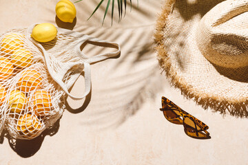 Summer flat lay on beige background. Straw hat, sunglasses, lemon fruits in eco friendly mesh shopping bag. Trendy palm shadow and sunlight, sun. Minimal summer travel fashion composition