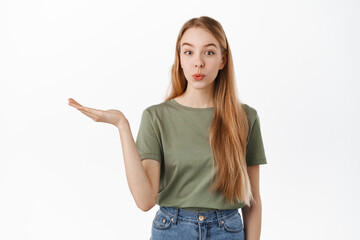 Fototapeta premium Cute blond girl holding empty hand, raising arm with opened palm as if display product, advertising at item, looking intrigued at camera, standing over white background