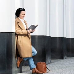 An adult woman fashionably dressed stands on the street near a building reading a book.