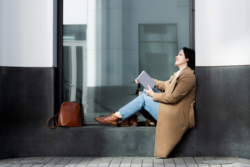 An adult woman fashionably dressed sits on the street on the windowsill of a building reading a book.