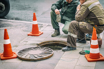 Workers sitting on over open sewer hatch on street near traffic cones. Repair of sewage,...