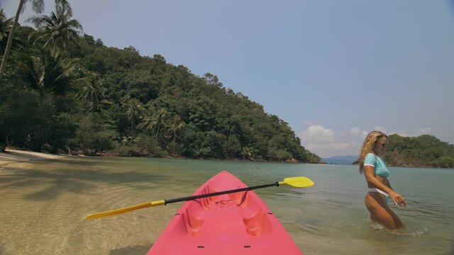Young woman in sunglasses pulls pink plastic canoe with paddle to azure sea water from beach with palms. The beginning of the boat trip on kayak, boarding sailing. Traveling to tropical countries.