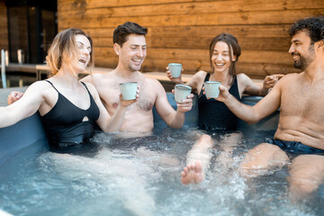 Company of friends steaming in a vat outdoors near the spa and lake. Women and man have fun and relax in wellness center. Raising glasses and cheers.