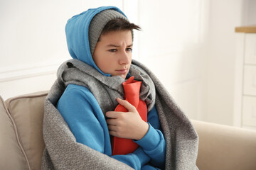 Sick teenage boy wrapped in blanket with hot water bottle on sofa at home