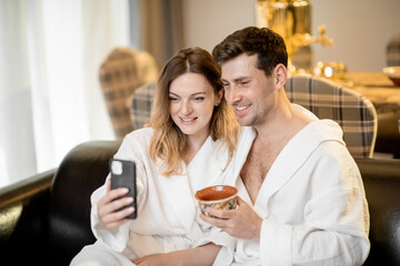 Woman and male in bathrobe sitting on sofa in living room of spa house and making a selfie. Relax and wellness concept. Couple spending and enjoy time together.