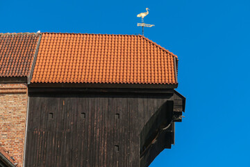 A close up on the rooftop of medieval port crane in Gdansk, Poland. The crane is made of dark wood. The rooftop is covered with tiles. A weather vane on top of the roof. Clear, blue sky above