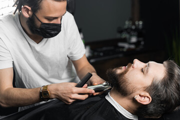 Beard shaving in barbershop. Barber with dreadlocks in black medical mask trim bearded man at quarantine coronavirus covid-19.