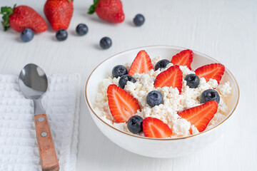 Quark or quarg - type of fresh dairy product made of fermented soured milk also called curd or cottage cheese served in plate with fresh strawberries and blueberries with spoon on white wooden table